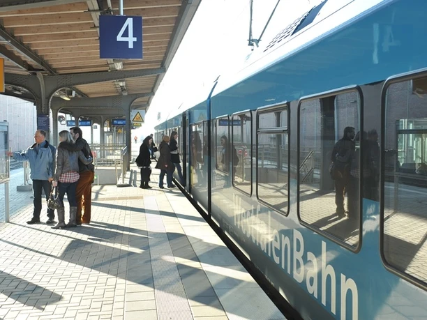 Menschen warten auf dem sonnigen Bahnsteig 4 in Paderborn, während ein Zug einfährt.
