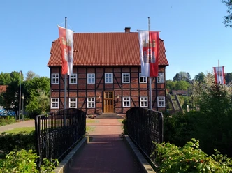 Spanckenhof Bad Wünnenberg Fachwerkhaus mit rotem Ziegeldach, umgeben von Bäumen. Zwei Flaggen weisen den Weg über eine Brücke.