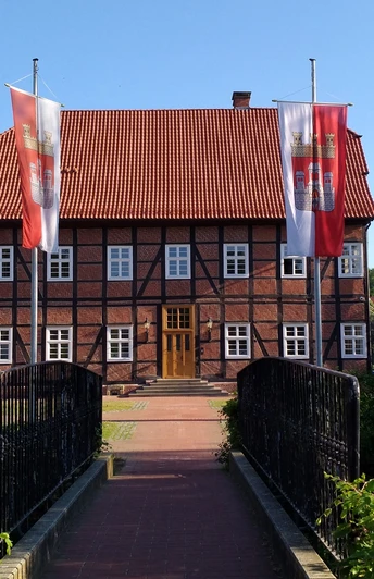 Spanckenhof Bad Wünnenberg Fachwerkhaus mit rotem Ziegeldach, umgeben von Bäumen. Zwei Flaggen weisen den Weg über eine Brücke.