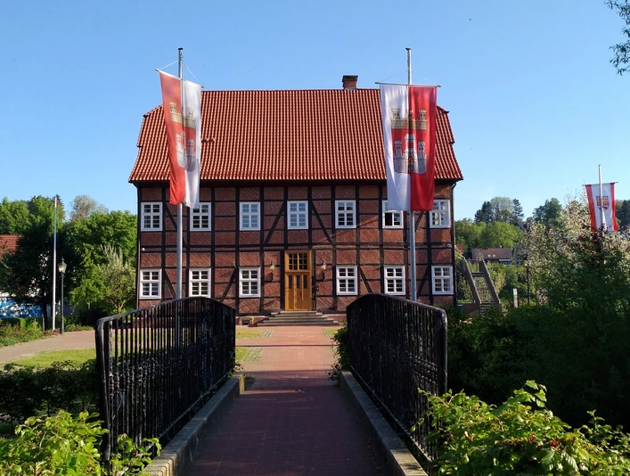 Spanckenhof Bad Wünnenberg Fachwerkhaus mit rotem Ziegeldach, umgeben von Bäumen. Zwei Flaggen weisen den Weg über eine Brücke.