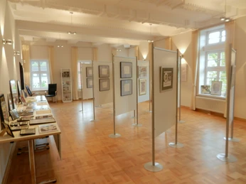 Ausstellung im Stucksaal Stucksaal mit hellen Wänden, Parkettboden und freistehenden Stellwänden mit gerahmten Kunstwerken.
