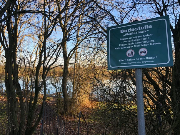 Badestelle am Wellier Kolk, von Bäumen umgeben, mit einem Warnschild, das Sicherheitsregeln erklärt.