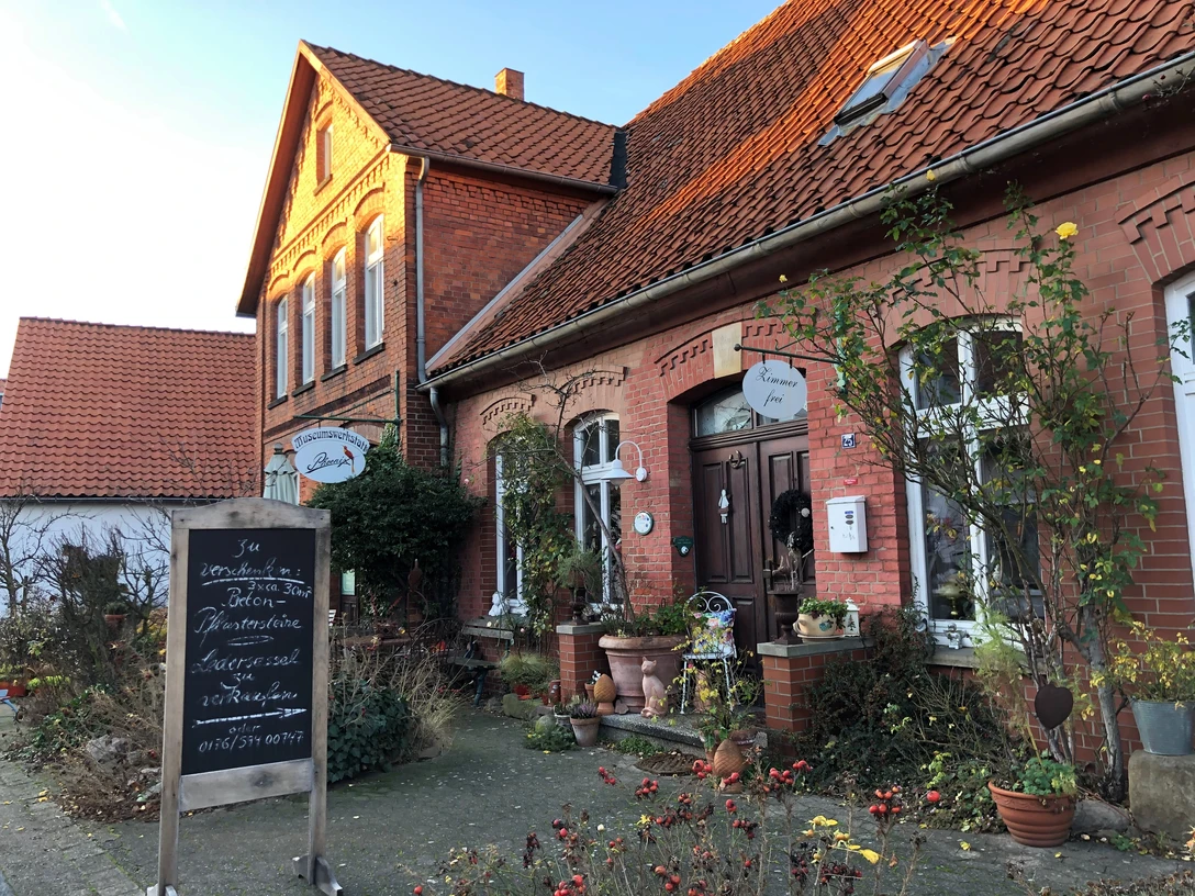 Museumswerkstatt Phönix Backsteingebäude mit Satteldach, Schildern am Eingang, Pflanzen auf Gehweg, sanftes Sonnenlicht.