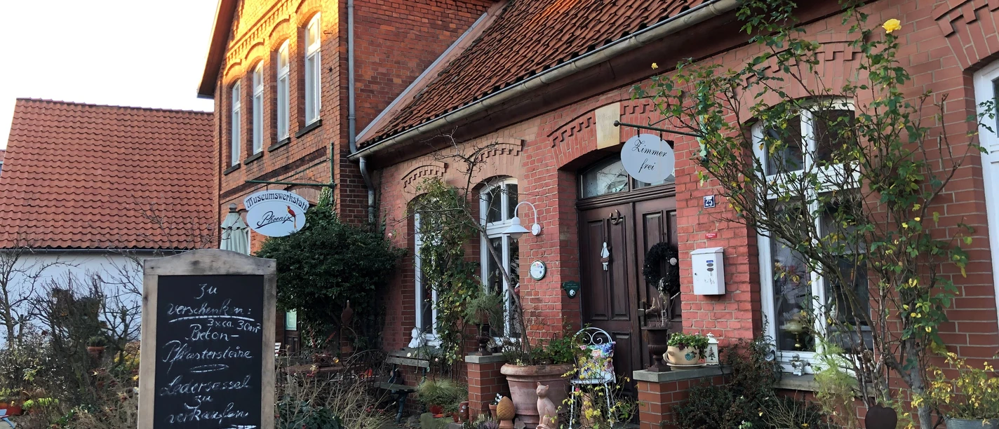 Museumswerkstatt Phönix Backsteingebäude mit Satteldach, Schildern am Eingang, Pflanzen auf Gehweg, sanftes Sonnenlicht.