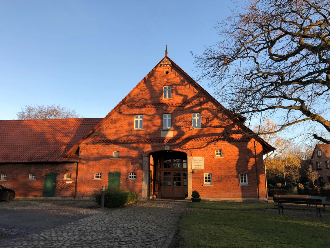 Mühlenbauhof Petershagen Historischer roter Backsteinbau mit steilem Dach, umgeben von Baum und gepflastertem Vorplatz.
