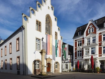 Rolandsäule vor historischem Gebäude auf einem belebten Marktplatz, flankiert von Bäumen und Häusern.