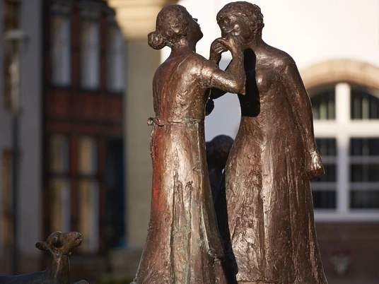 Bronzeskulptur zweier Frauen, die einander ins Ohr flüstern, im frühen Morgenlicht am Marktplatz.