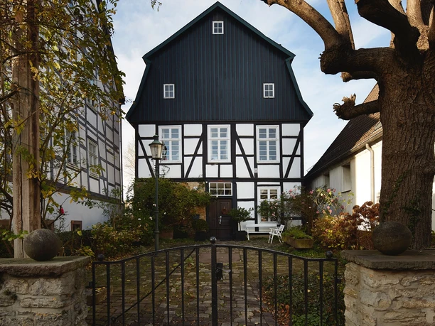 Ein historisches Fachwerkhaus mit dunklem Giebel, umgeben von Bäumen, vor einem schmiedeeisernen Tor.