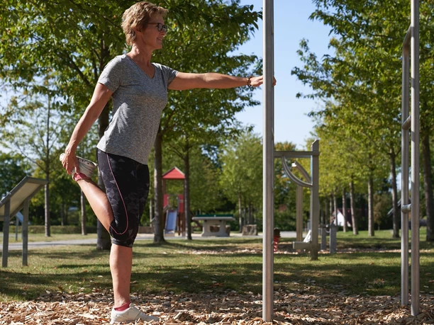 Generationenpark Frau dehnt sich im Grünen an einem Fitnessgerät; im Hintergrund Parkausstattung und blauer Himmel.