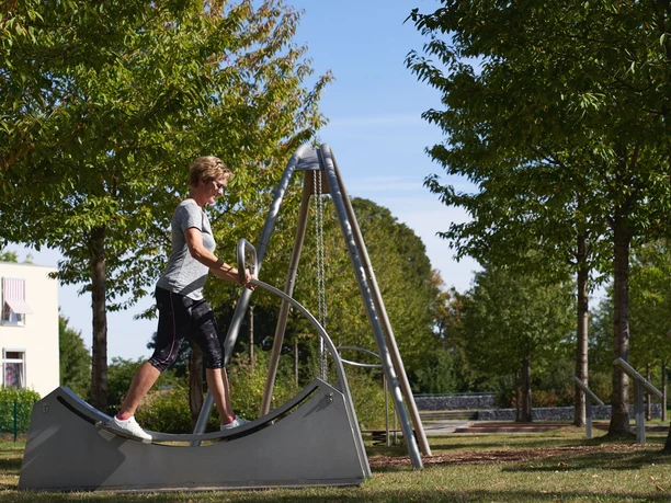 Generationenpark Ein Junge balanciert auf einem modernen Spielgerät im Grünen eines Parks unter klarem, blauem Himmel.