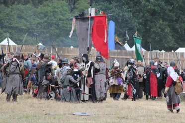 Conquest of Mythodea Menschen in mittelalterlichen Kostümen versammeln sich auf einer Wiese vor Holzzäunen und bunten Flaggen.