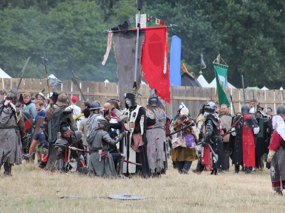 Conquest of Mythodea Menschen in mittelalterlichen Kostümen versammeln sich auf einer Wiese vor Holzzäunen und bunten Flaggen.People in medieval costumes gather on a meadow in front of wooden fences and colorful flags.Folk i middelalderdragter samles på en eng foran træhegn og farverige flag.Mensen in middeleeuwse kostuums verzamelen zich op een weiland voor houten hekken en kleurrijke vlaggen.