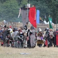 Conquest of Mythodea Menschen in mittelalterlichen Kostümen versammeln sich auf einer Wiese vor Holzzäunen und bunten Flaggen.