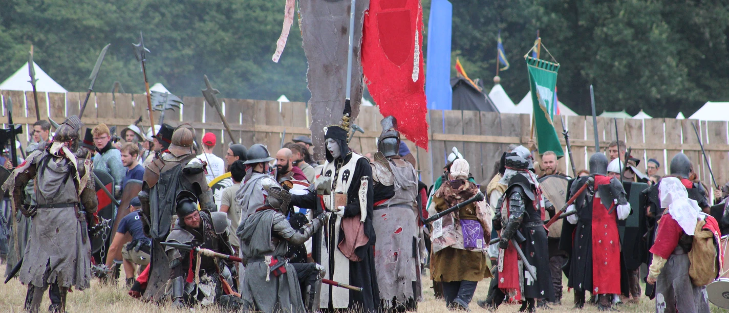 Conquest of Mythodea People in medieval costumes gather on a meadow in front of wooden fences and colorful flags.