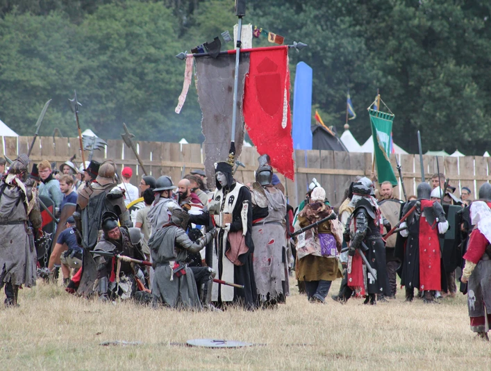 Conquest of Mythodea Menschen in mittelalterlichen Kostümen versammeln sich auf einer Wiese vor Holzzäunen und bunten Flaggen.People in medieval costumes gather on a meadow in front of wooden fences and colorful flags.Folk i middelalderdragter samles på en eng foran træhegn og farverige flag.Mensen in middeleeuwse kostuums verzamelen zich op een weiland voor houten hekken en kleurrijke vlaggen.