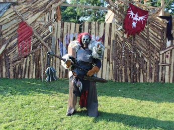 Conquest of Mythodea Ein Krieger in Rüstung steht bewaffnet mit einer großen Axt vor einem rustikalen Holzverhau im Freien.A warrior in armor stands armed with a large axe in front of a rustic wooden hut outside.En kriger i rustning står bevæbnet med en stor økse foran en rustik træhytte udenfor.Een krijger in harnas staat gewapend met een grote bijl voor een rustieke houten hut buiten.