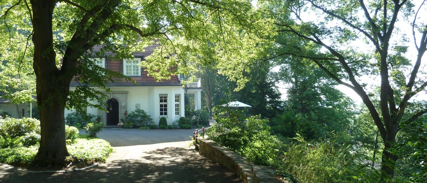 Haus Hünenburg in Achim-Baden, umgeben von grünen Bäumen, lässt Sonnenlicht durch das Laub scheinen.