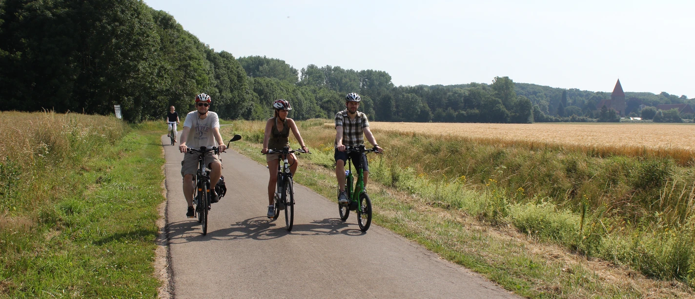 Radfahren Drei Radfahrer auf einem asphaltierten Weg in einer ländlichen Landschaft mit Feldern und Bäumen.