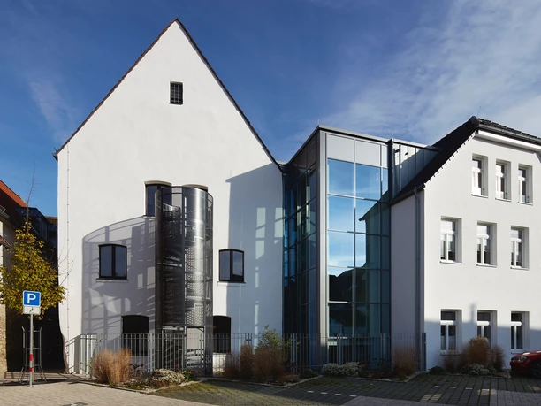 Weiße Gebäudefassade mit moderner Glasfront und Zylinderanbau unter blauem Himmel.