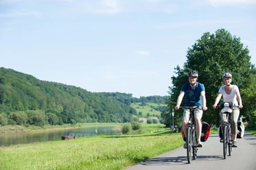 Radfahrer Wehrden Weserradweg Radfahrer auf einem Fahrradweg entlang eines Flusses, umgeben von grünen Hügeln und Bäumen im Sommer.