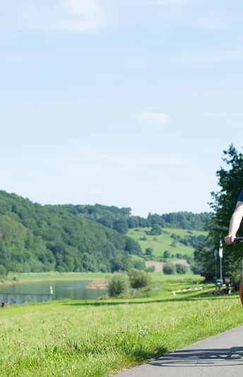 Radfahrer Wehrden Weserradweg Radfahrer auf einem Fahrradweg entlang eines Flusses, umgeben von grünen Hügeln und Bäumen im Sommer.