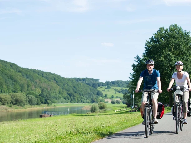 Radfahrer Wehrden Weserradweg Radfahrer auf einem Fahrradweg entlang eines Flusses, umgeben von grünen Hügeln und Bäumen im Sommer.