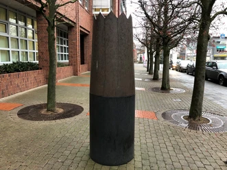 Große Skulptur einer Holz- und Metallkrone auf Pflasterweg, flankiert von Bäumen und Backsteingebäude.