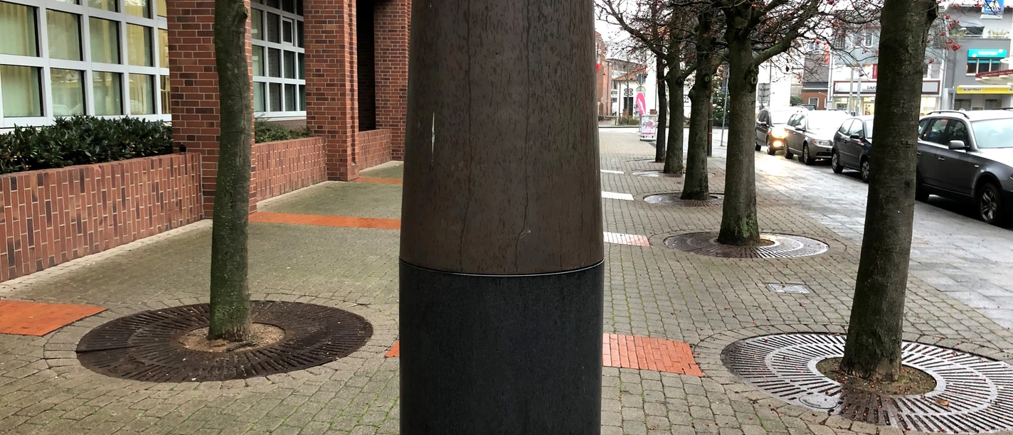 Große Skulptur einer Holz- und Metallkrone auf Pflasterweg, flankiert von Bäumen und Backsteingebäude.