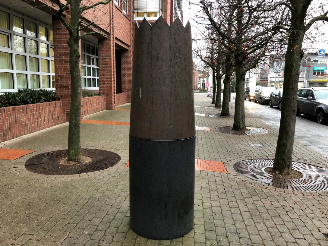 Skulptur Krone Große Skulptur einer Holz- und Metallkrone auf Pflasterweg, flankiert von Bäumen und Backsteingebäude.