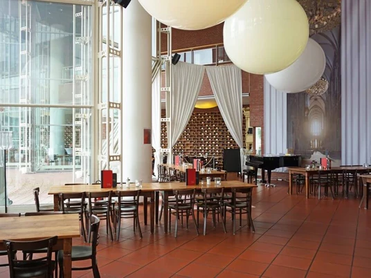 Café Restaurant Ludwig im Museum Großzügiger Restaurant Innenbereich mit bodenlanger Glasfront. Dunkle Holztische erschaffen eine Gemütlichkeit und an der Decke hängen riesige ballonähnliche Lampen.Spacious restaurant interior with floor-to-ceiling glass front. Dark wooden tables create a cozy atmosphere and huge balloon-like lamps hang from the ceiling.