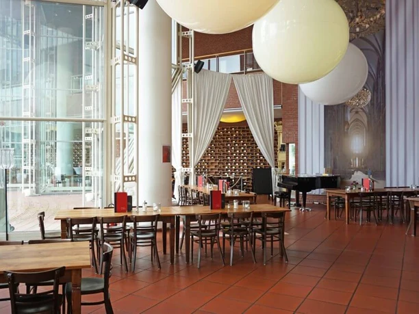 Café Restaurant Ludwig im Museum Großzügiger Restaurant Innenbereich mit bodenlanger Glasfront. Dunkle Holztische erschaffen eine Gemütlichkeit und an der Decke hängen riesige ballonähnliche Lampen.