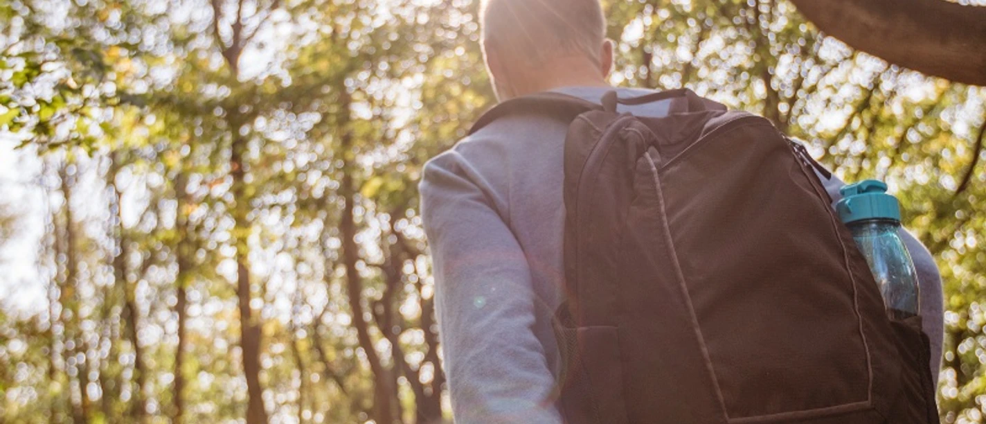 Wandern Ein Wanderer mit Rucksack und Karte spaziert entlang eines Waldpfades im Sonnenlicht.