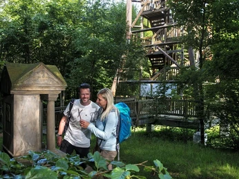 Ein Paar steht neben einem historischen Bildstock, umgeben von üppigem Grün und einem Holzturm.