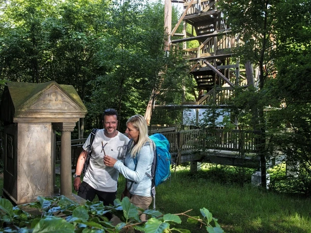 Ein Paar steht neben einem historischen Bildstock, umgeben von üppigem Grün und einem Holzturm.