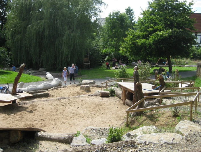 Ein Spielplatz mit Sandfläche, Holzskulpturen und Bäumen im Hintergrund, Kinder spielen fröhlich.