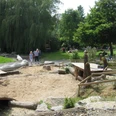 image Ein Spielplatz mit Sandfläche, Holzskulpturen und Bäumen im Hintergrund, Kinder spielen fröhlich.