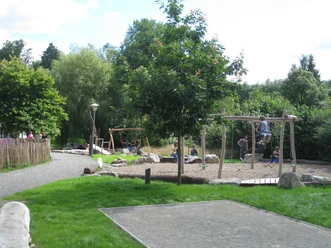 Spielplatz mit Holzschaukeln und grünem Umfeld; Kinder beim Spielen an einem sonnigen Tag im Freien.