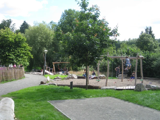 image Spielplatz mit Holzschaukeln und grünem Umfeld; Kinder beim Spielen an einem sonnigen Tag im Freien.
