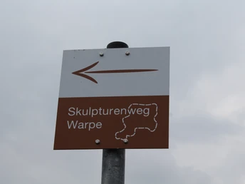Schild Skulturenweg Warpe Wegweiser mit Pfeil und Aufschrift "Skulpturenweg Warpe" vor bewölktem Himmel bei GPS-Koordinaten.