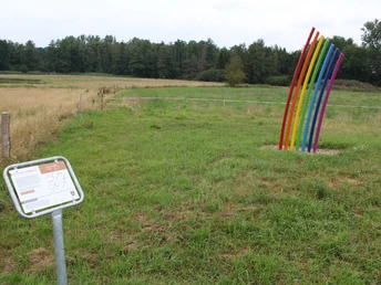 Bunte Skulptur in Regenbogenform auf einer grünen Wiese in einer ländlichen Umgebung aufgestellt.