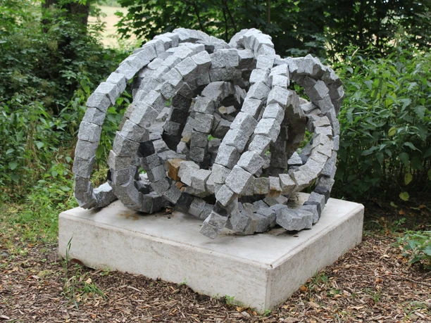 Skulptur Rolle vorwärts Een abstracte sculptuur van verstrengelde grijze granieten blokken staat op een betonnen sokkel in het groen.