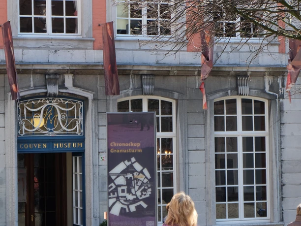 Aachener Chronoskop Museum.JPG Fassade des Couven Museums. Eine Person betrachtet ein Informationsdisplay vor dem Eingang. Das Gebäude besticht durch große Fenster und eine orangefarbene Fassade.