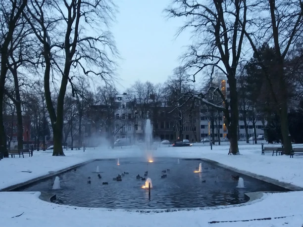 Der verschneite Kurpark zeigt einen Teich mit Fontänen, umgeben von schneebedeckten Bäumen.