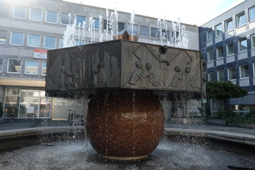 Runder Sockel mit verzierten Platten, aus denen Wasserfontänen empor schießen.