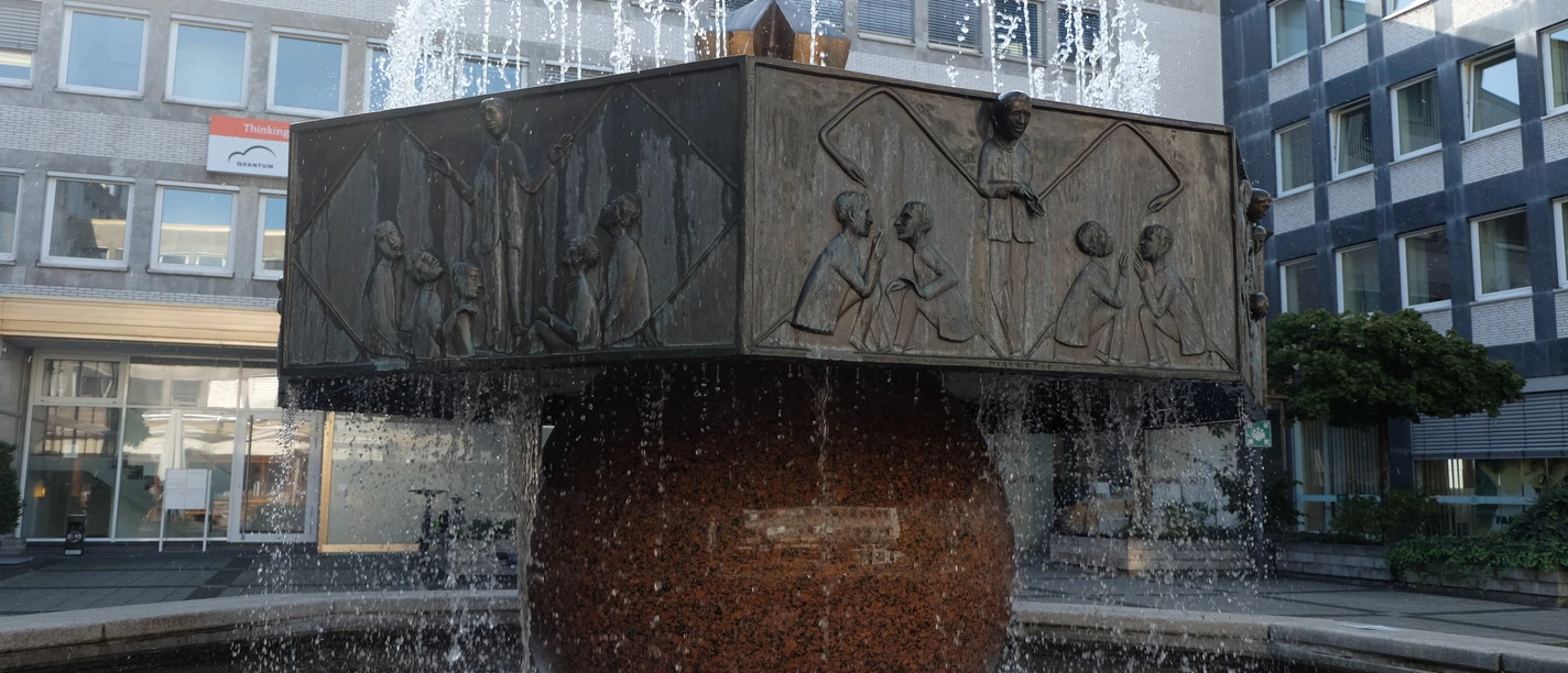 Karlshofbrunnen Runder Sockel mit verzierten Platten, aus denen Wasserfontänen empor schießen.