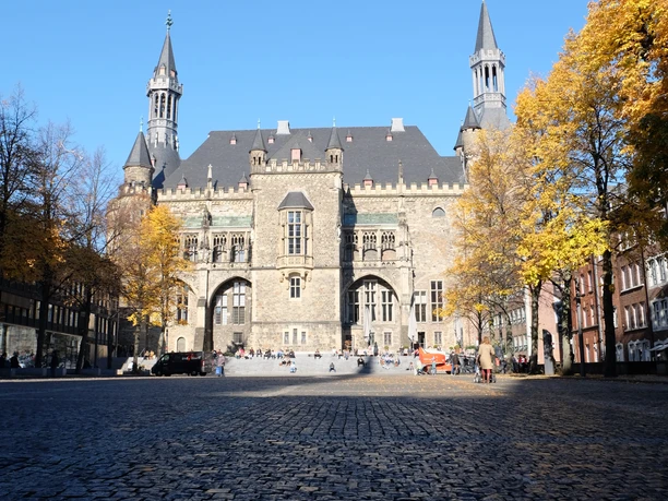 Rathaus mit bunten Laubbäumen unter blauem Himmel und auf Kopfsteinpflaster.