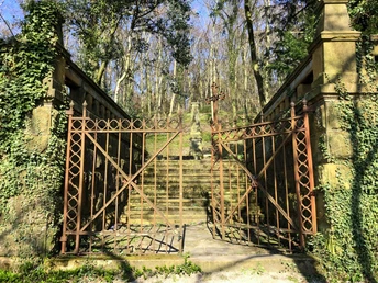 'Eingang zum Waldbegräbnis Vogelsang Historisches Metalltor führt zu einer von Bäumen umrahmten steinernen Treppe in einem bewaldeten Park.