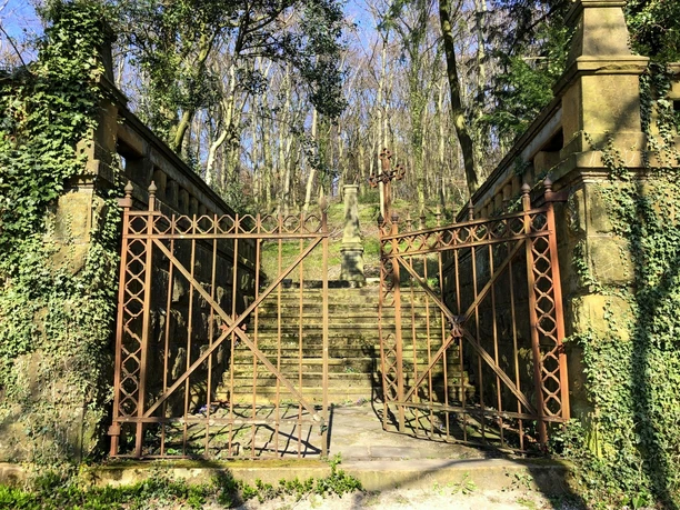'Eingang zum Waldbegräbnis Vogelsang Historisches Metalltor führt zu einer von Bäumen umrahmten steinernen Treppe in einem bewaldeten Park.