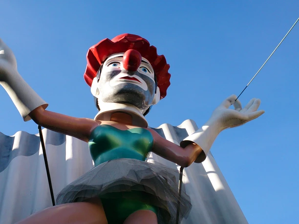 Eine fröhliche Clownsfigur in farbenfrohem Kostüm und roter Nase, die ihre Arme zur Begrüßung weit ausstreckt.