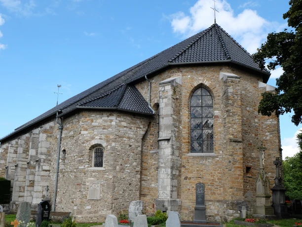 St. Stephanus Kirche aus hellbraunen Steinen vor einer Grabesstätte unter blauem Himmel.